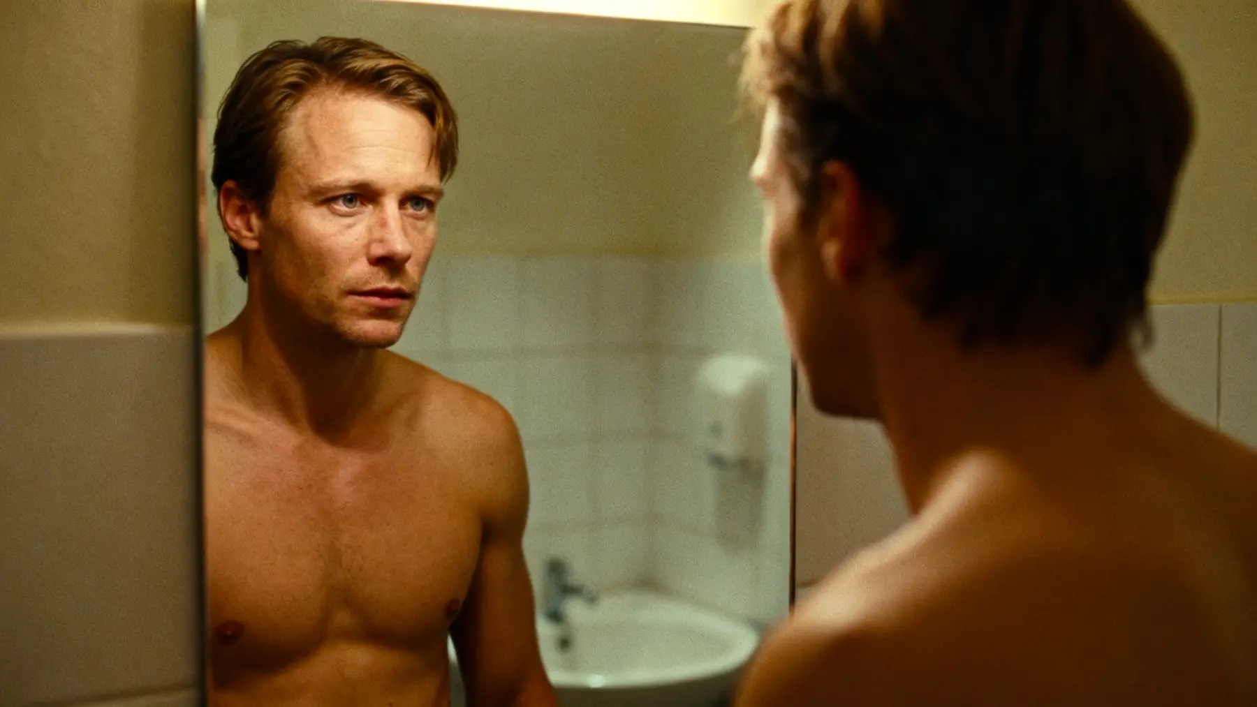Shirtless man studying his own reflection in a bathroom mirror