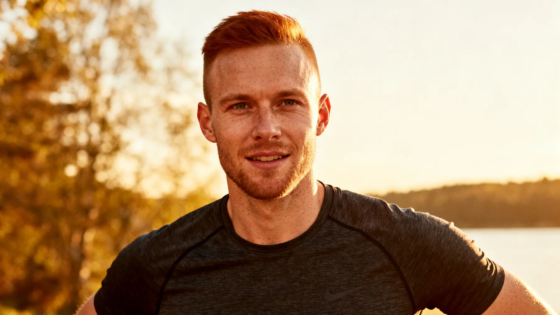 Fit red-haired man in an athletic shirt smiling by a lake at sunset