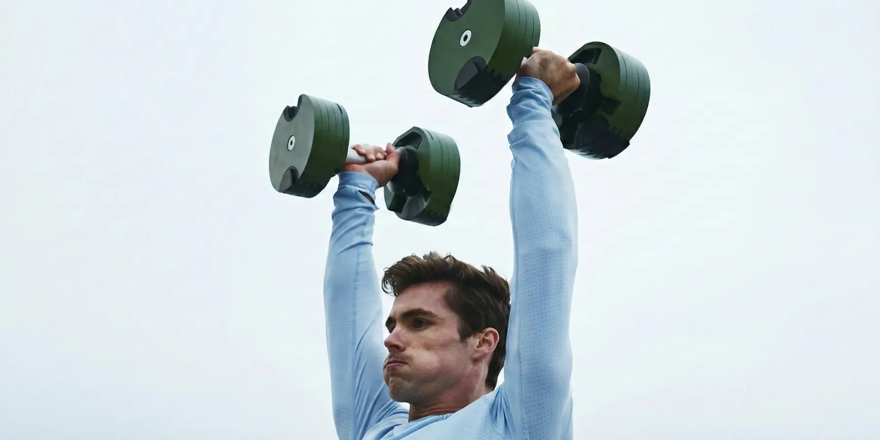Man pressing dumbbells overhead outdoors against a pale grey sky