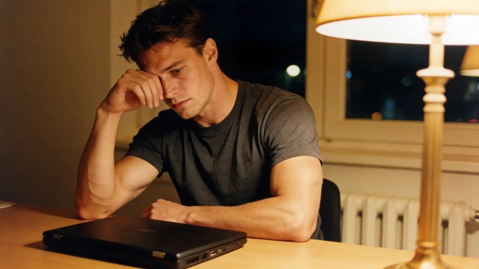 Tired young man pushing back from a closed laptop, hand on his face