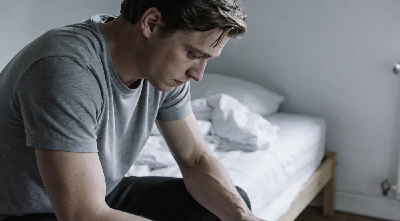 A young man sitting heavily on the edge of his bed in flat grey light, low.