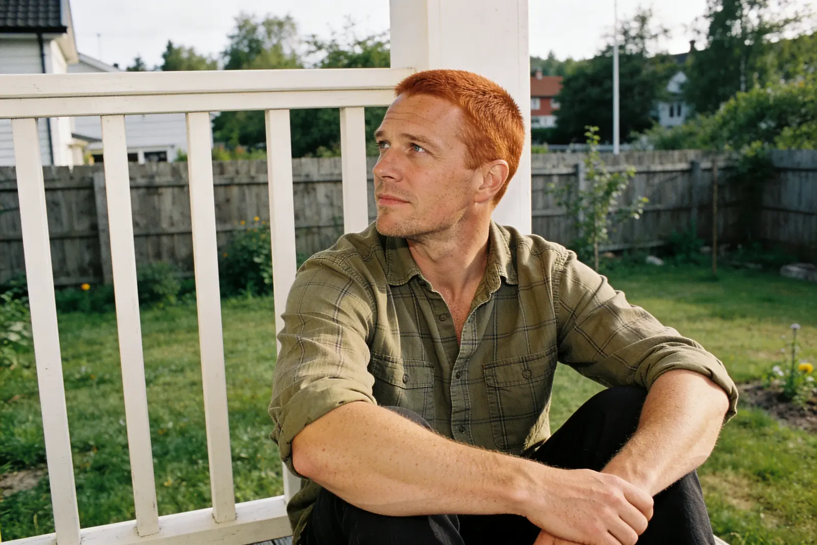 Red-haired man sitting on a porch step gazing away, pensive, in a green backyard