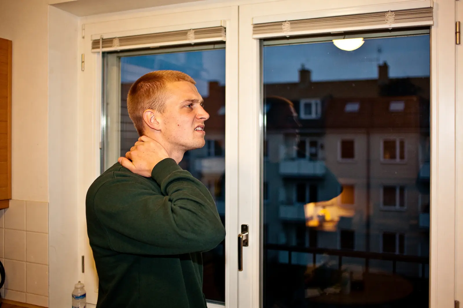 Man in a dark sweater rubbing his neck by a window at dusk, looking strained