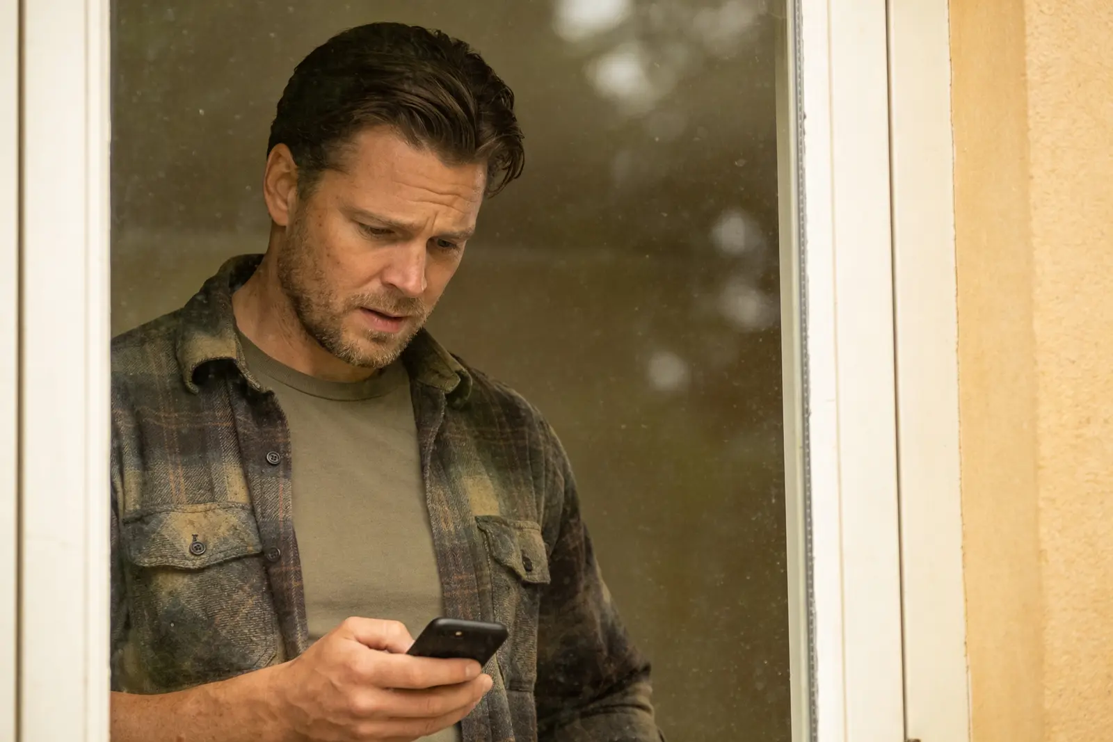 Man in a flannel shirt frowning at his phone in a doorway