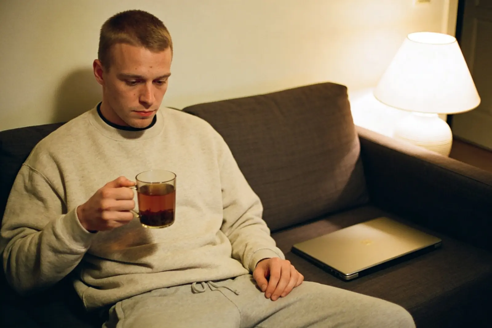 Young man in gray sweats sipping tea on a couch beside a closed laptop