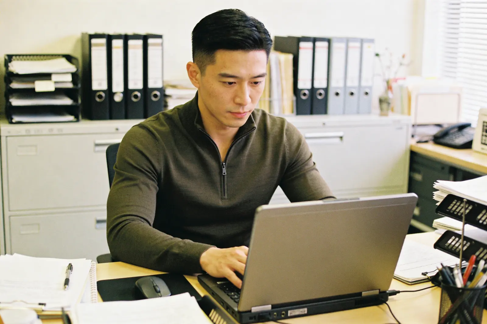 Focused man working on a laptop at a cluttered office desk with binders