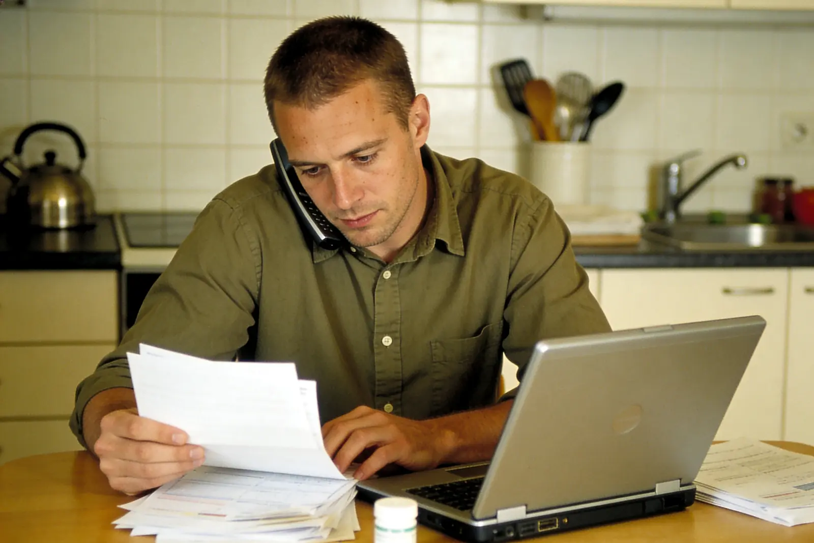 Man on a phone reviewing paperwork at a kitchen table beside a laptop