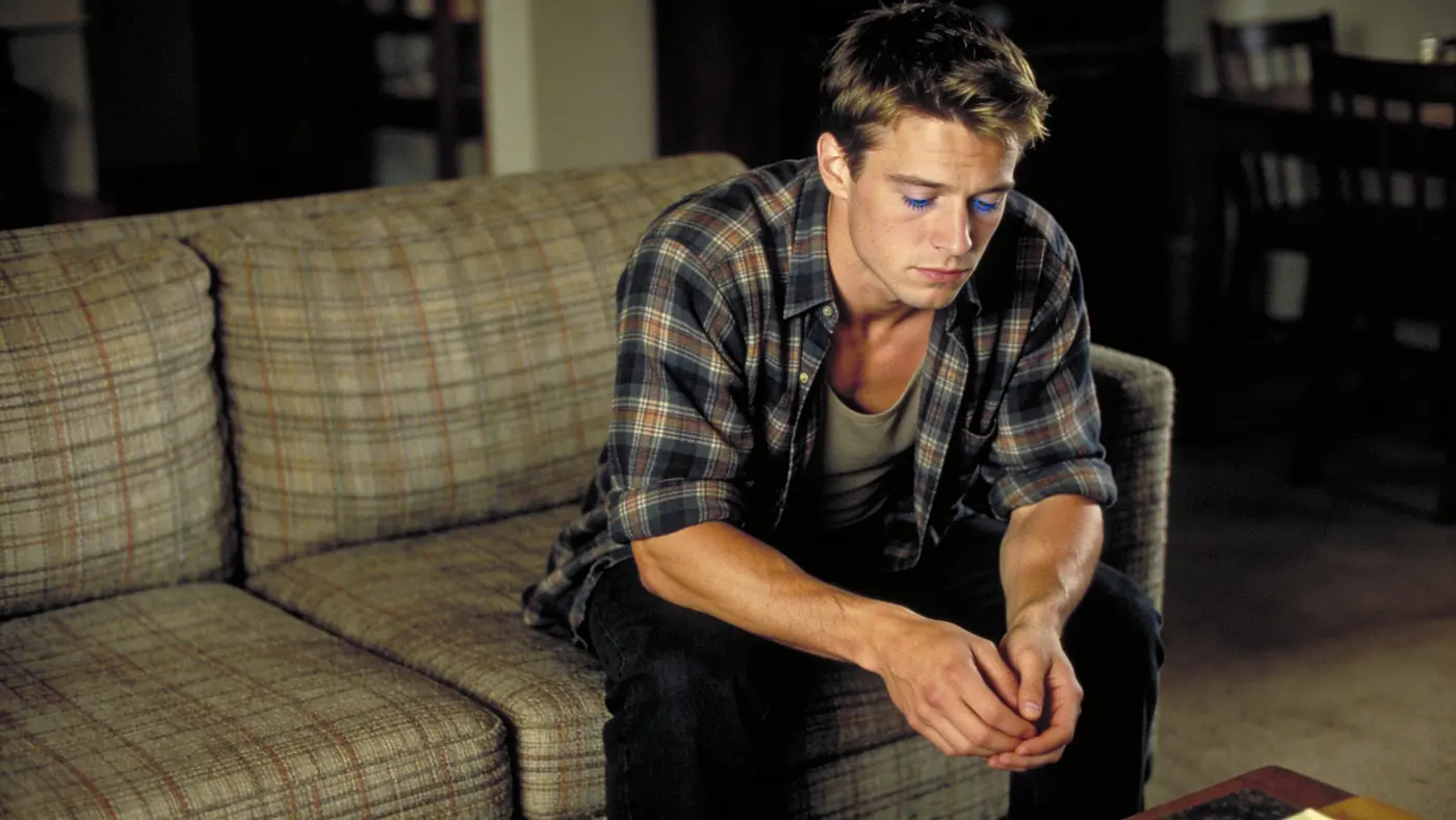 A young man sits on the edge of a couch with head lowered and eyes closed, worn down by a side effect.