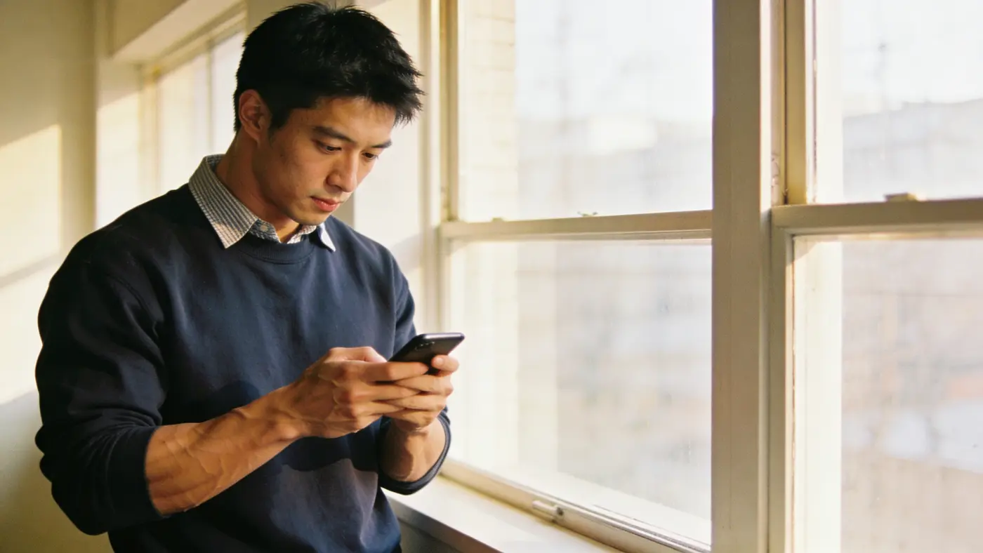 A young man at a bright window glances at his phone with returning focus, re-engaging with the day.