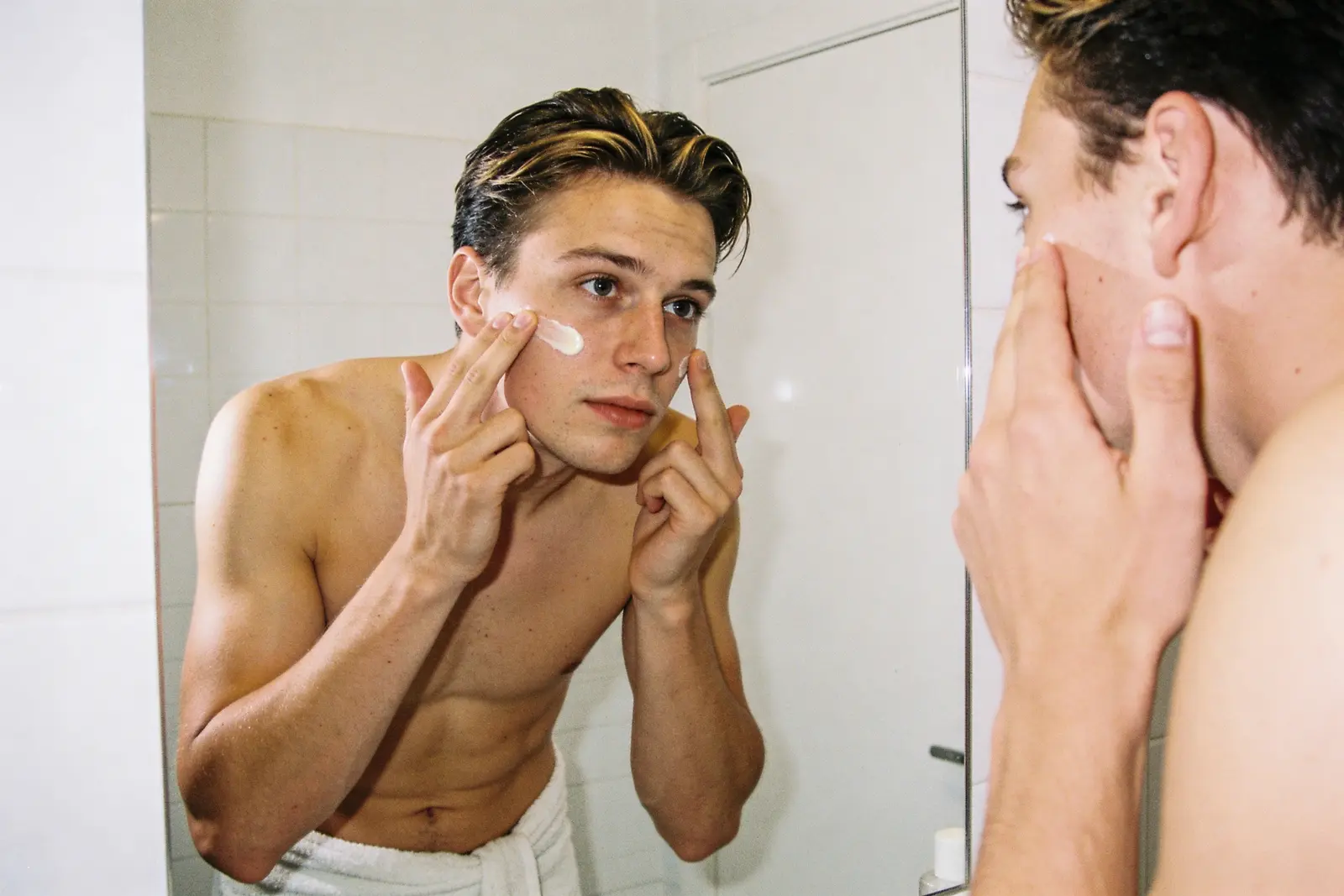 Shirtless young man applying moisturizer to his face at a bathroom mirror