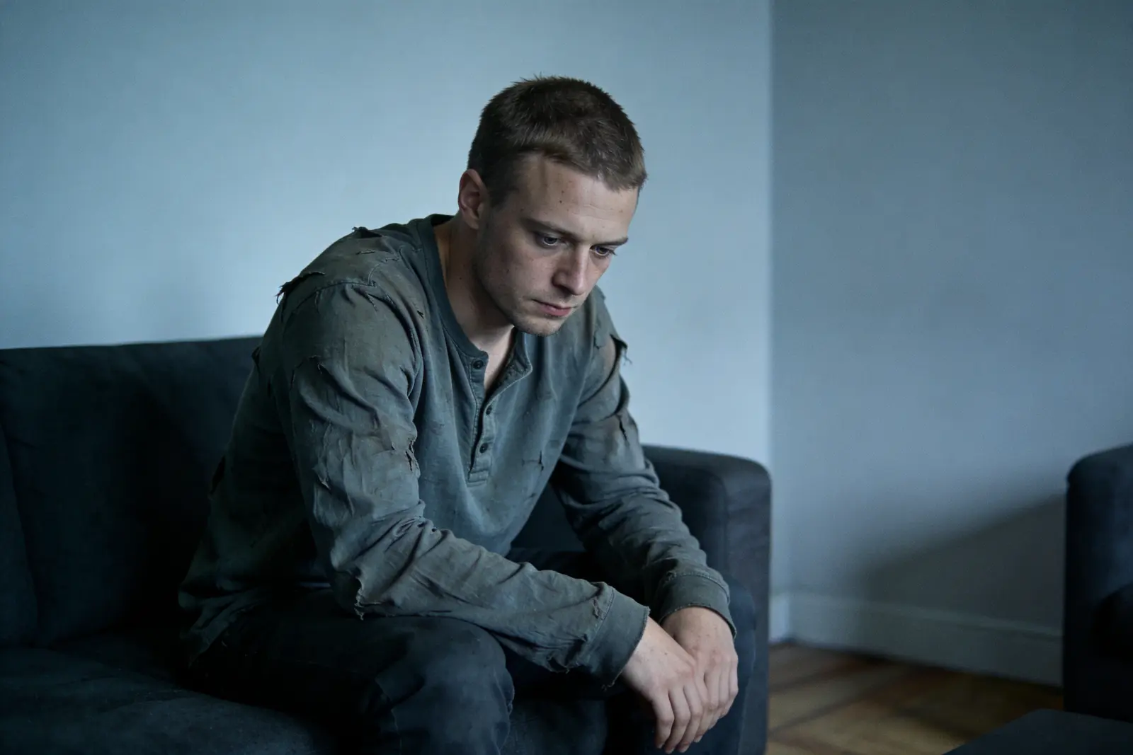 Young man in his late twenties sitting heavily on the couch edge in flat grey daylight