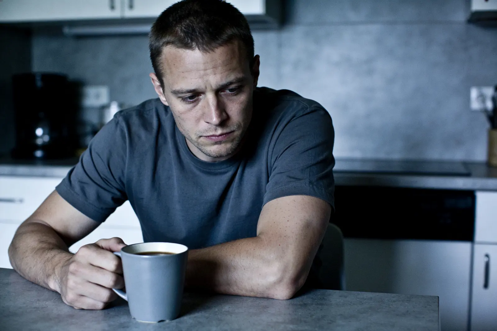 Exhausted man slumped at the kitchen table at dawn, coffee untouched