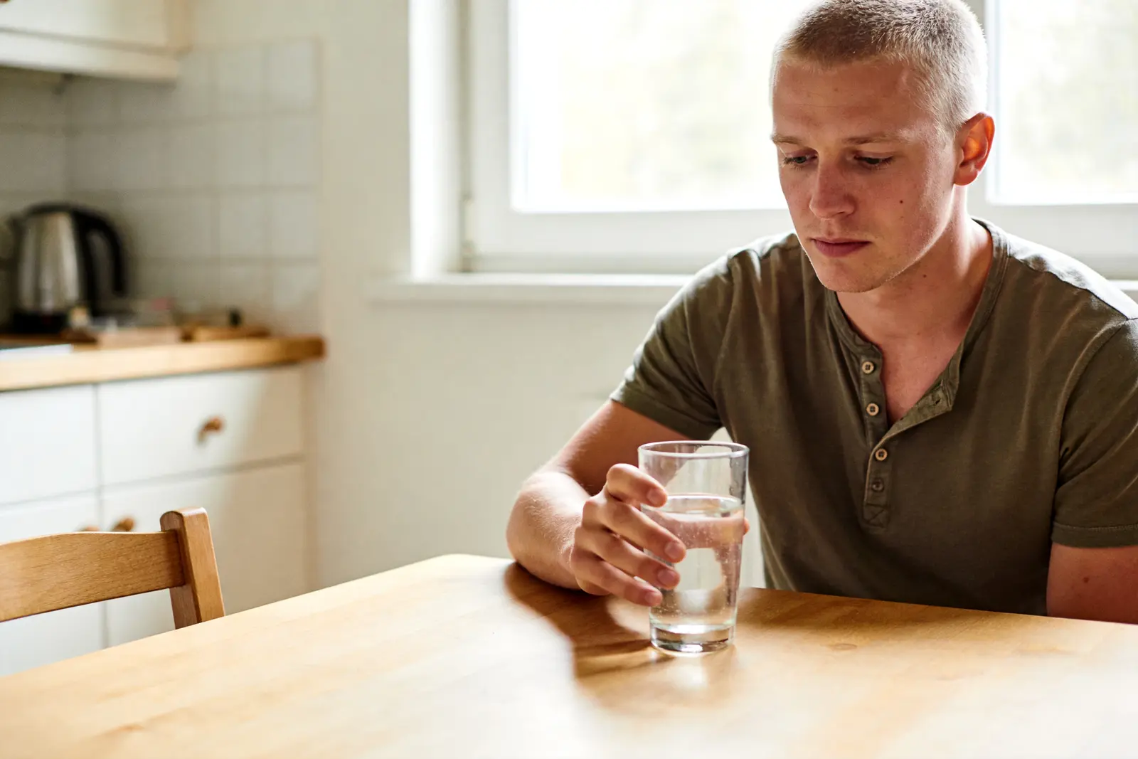 Man sitting calmly at a kitchen table in morning light, holding a glass of water
