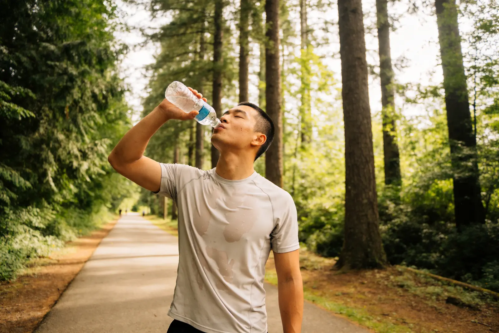 Man pausing during a run to drink water from a bottle on a tree-lined path