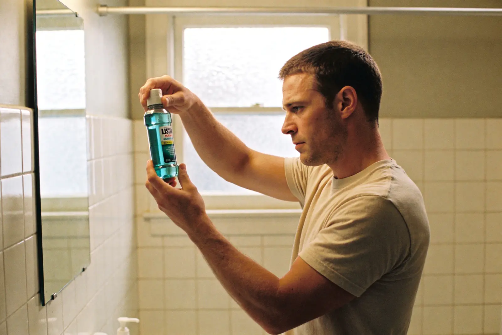 Man at the bathroom mirror reading the label on a bottle of mouthwash