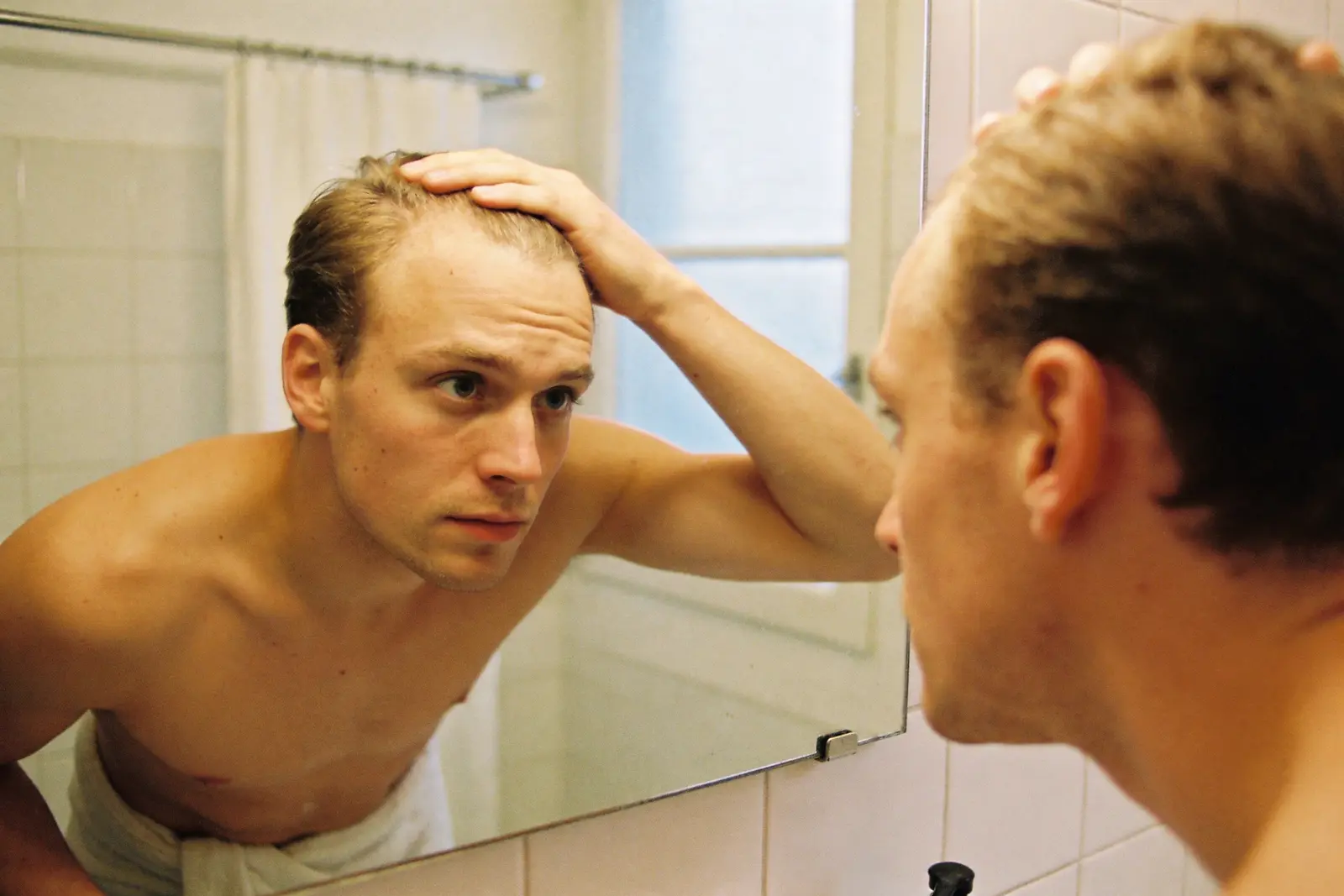 Young man checking his hairline in a bathroom mirror under morning light