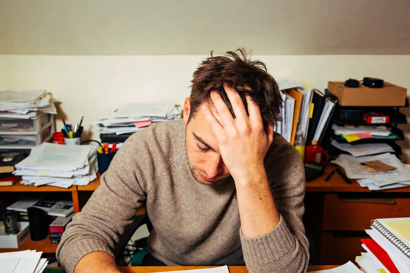 Man at a cluttered desk rubbing his eyes, overwhelmed by a small setback